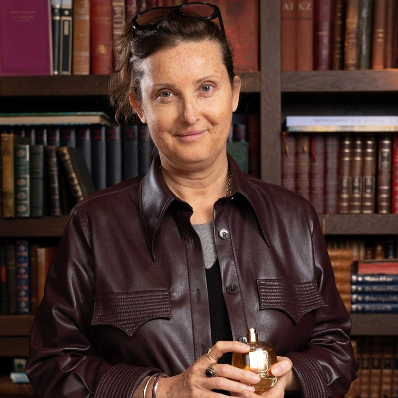 Cécile Vialla, oprichter en parfumeur van Maison de Parfum BERRY, gefotografeerd voor een boekenkast terwijl zij een parfumflacon vasthoudt.
