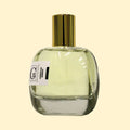 A studio product photograph of a clear glass perfume bottle filled with pale yellow-green fragrance, topped with a reflective gold cylindrical cap and set against a soft cream background. A white label on the front displays the Collection Trente-Cinq logo and the name “Annalia Débat.” The transparent bottle showcases the light-colored perfume, while subtle reflections on the glass emphasize its rounded, sculptural form.