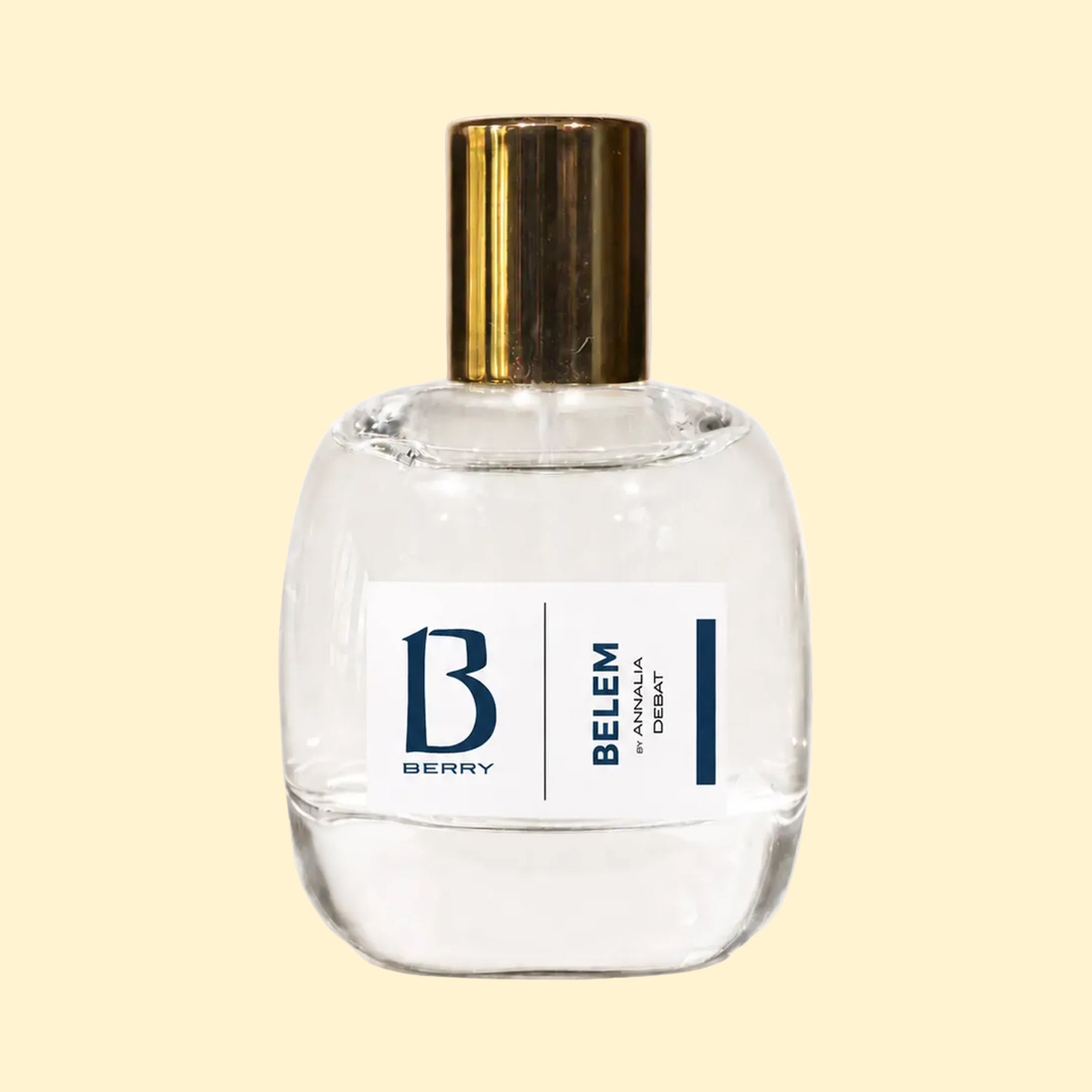 A studio product photograph of a clear glass perfume bottle filled with pale yellow-green fragrance, topped with a reflective gold cylindrical cap and set against a soft cream background. A white label on the front displays the Parfums Berry logo and the name “Annalia Débat.” The transparent bottle showcases the light-coloured perfume, while subtle reflections on the glass emphasise its rounded, sculptural form.