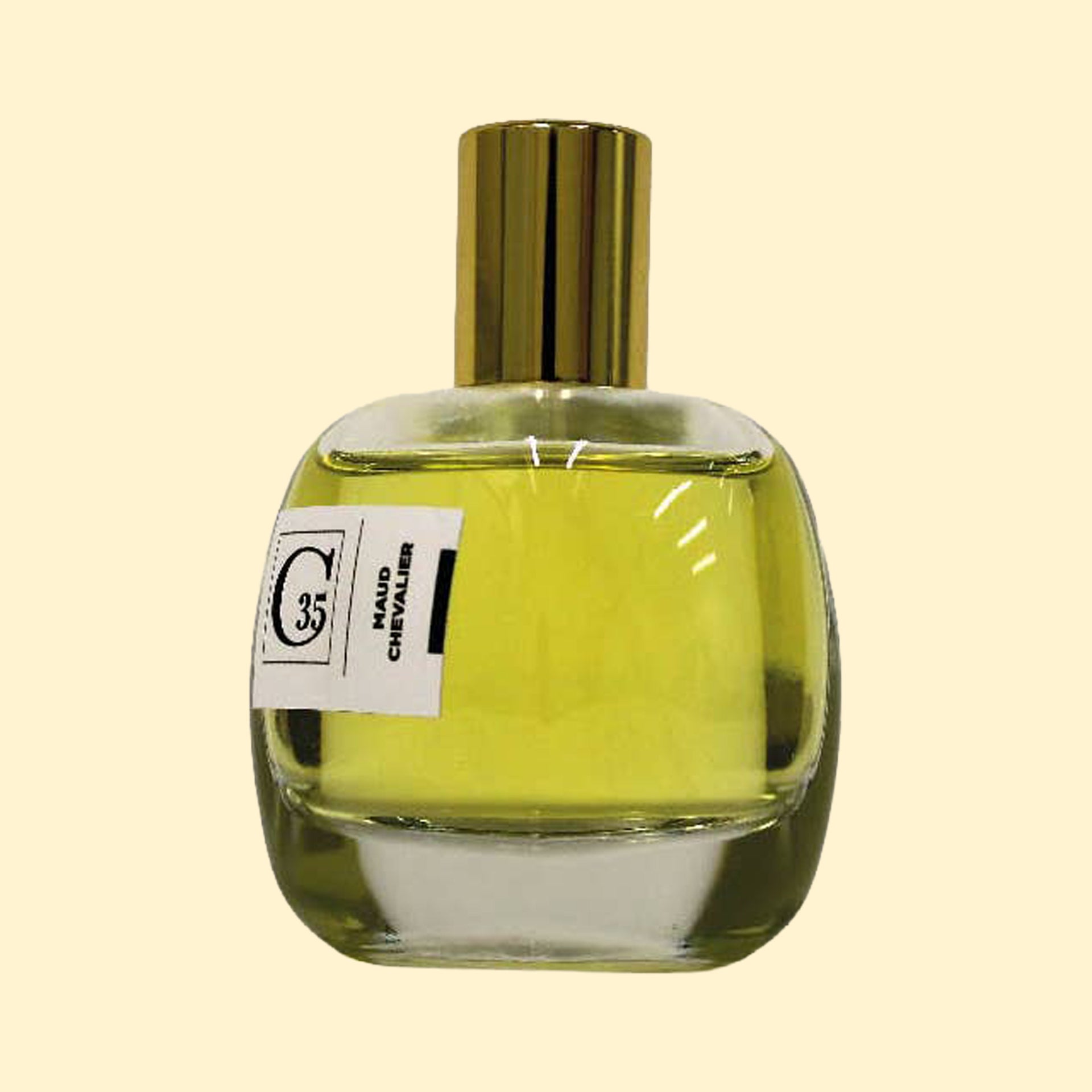 A studio product photograph of a clear glass perfume bottle filled with yellow-green fragrance, topped with a reflective gold cylindrical cap and set against a soft cream background. A white label on the front displays the Collection Trente-Cinq logo and the name “Maud Chevalier.” The transparent bottle reveals the luminous color of the perfume, while soft reflections on the glass emphasize its rounded, sculptural shape.