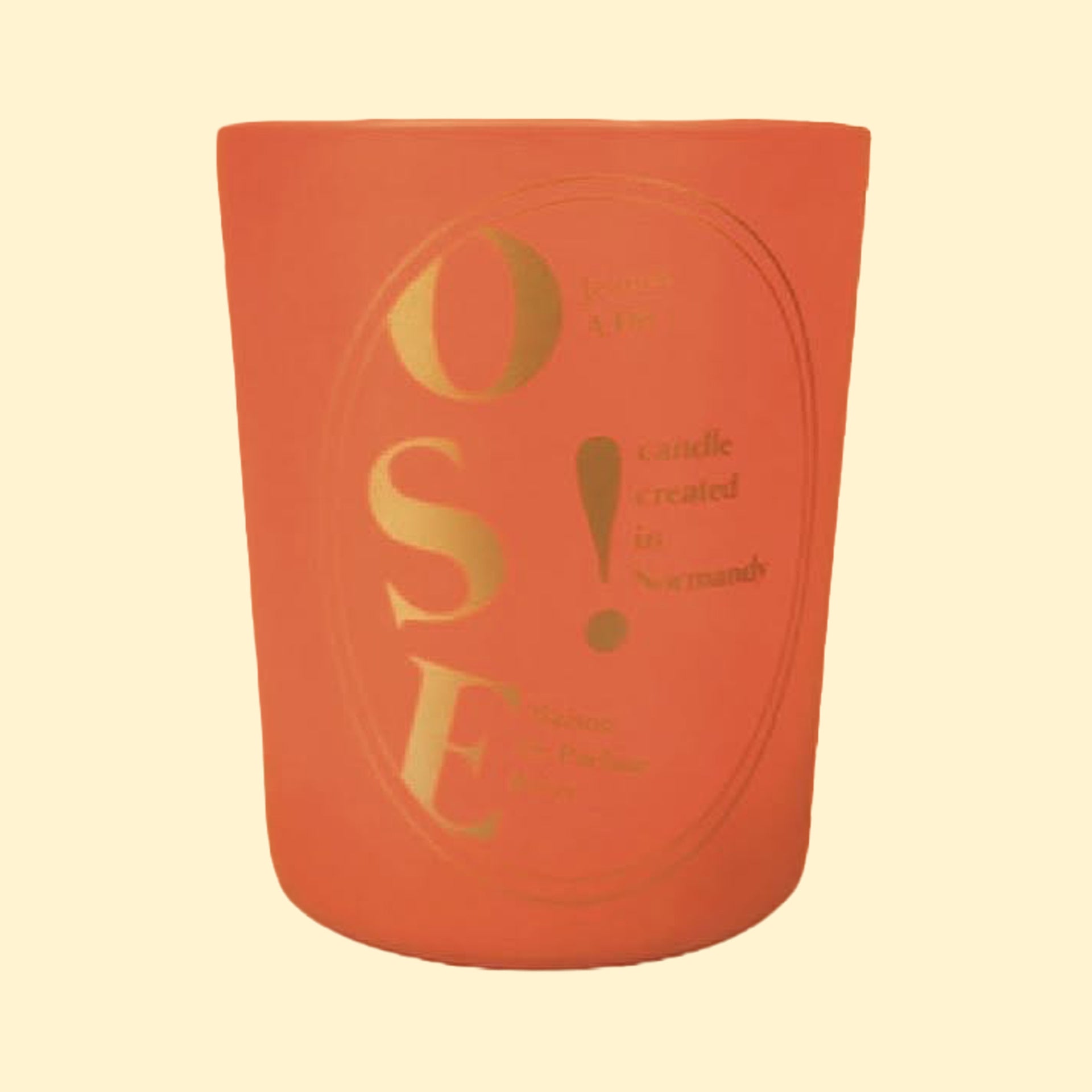A studio product photograph of a matte terracotta-orange candle vessel set against a soft cream background. The front features gold typography with the large letters “OSE!” and the phrase “candles created in Normandy.” The minimalist cylindrical container has a warm, contemporary appearance, with metallic lettering that contrasts against the rich orange surface.