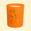 A studio product photograph of a matte terracotta-orange candle vessel set against a soft cream background. The front features gold typography with the large letters “OSE!” and the phrase “candle created in Normandy.” The minimalist cylindrical container has a warm, contemporary appearance, with metallic lettering that contrasts against the rich orange surface.