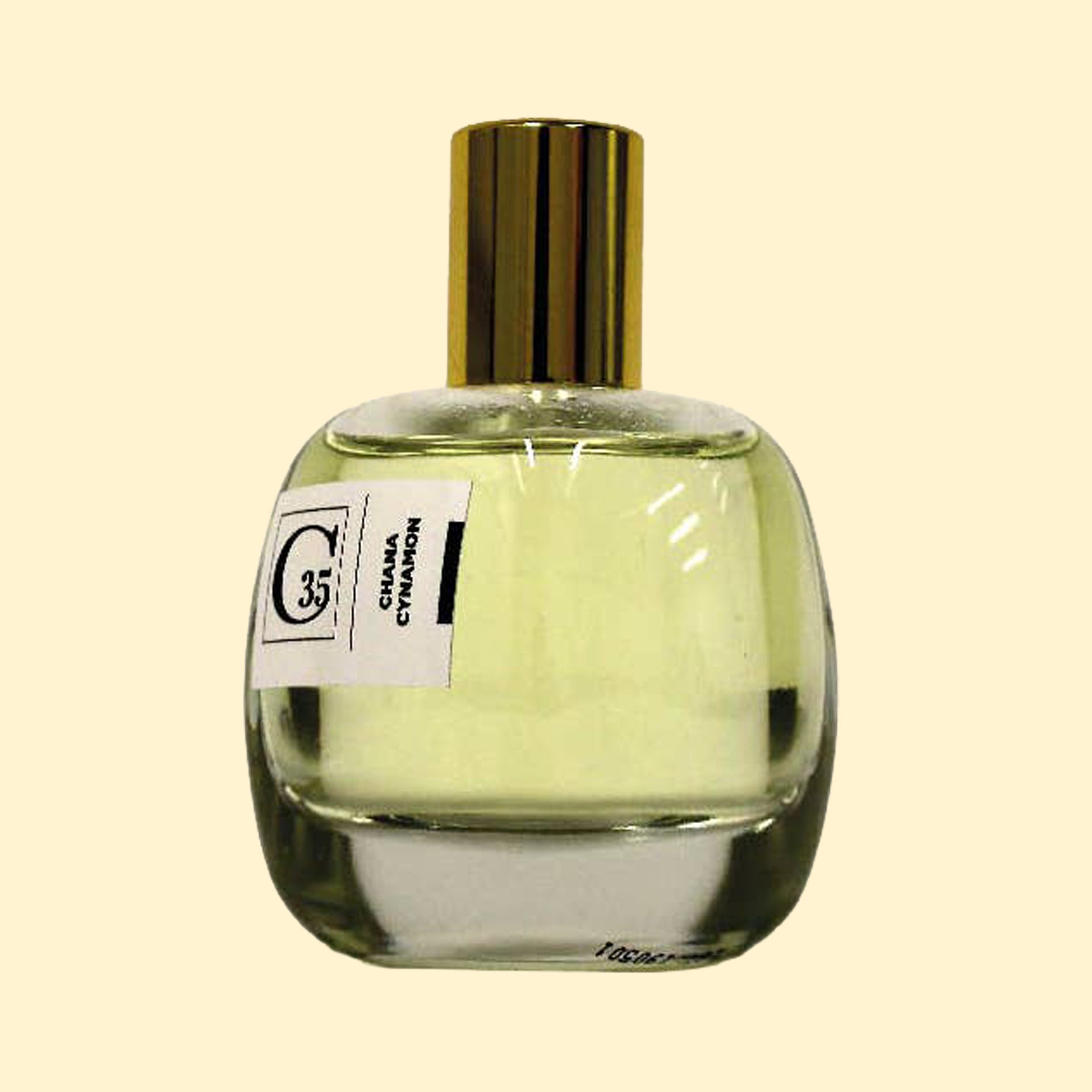 A studio product photograph of a clear glass perfume bottle filled with pale yellow-green fragrance, topped with a reflective gold cylindrical cap and set against a soft cream background. A white label on the front displays the Collection Trente-Cinq logo and the name “Chana Cynamon.” The transparent bottle showcases the light-colored perfume, while soft reflections on the glass emphasize its rounded, sculptural silhouette.
