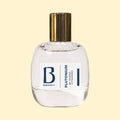 A studio product photograph of a clear glass perfume bottle filled with pale yellow-green fragrance, topped with a reflective gold cylindrical cap and set against a soft cream background. A white label on the front displays the Parfums Berry logo and the name “Chana Cynamon.” The transparent bottle showcases the light-coloured perfume, while soft reflections on the glass emphasise its rounded, sculptural silhouette.