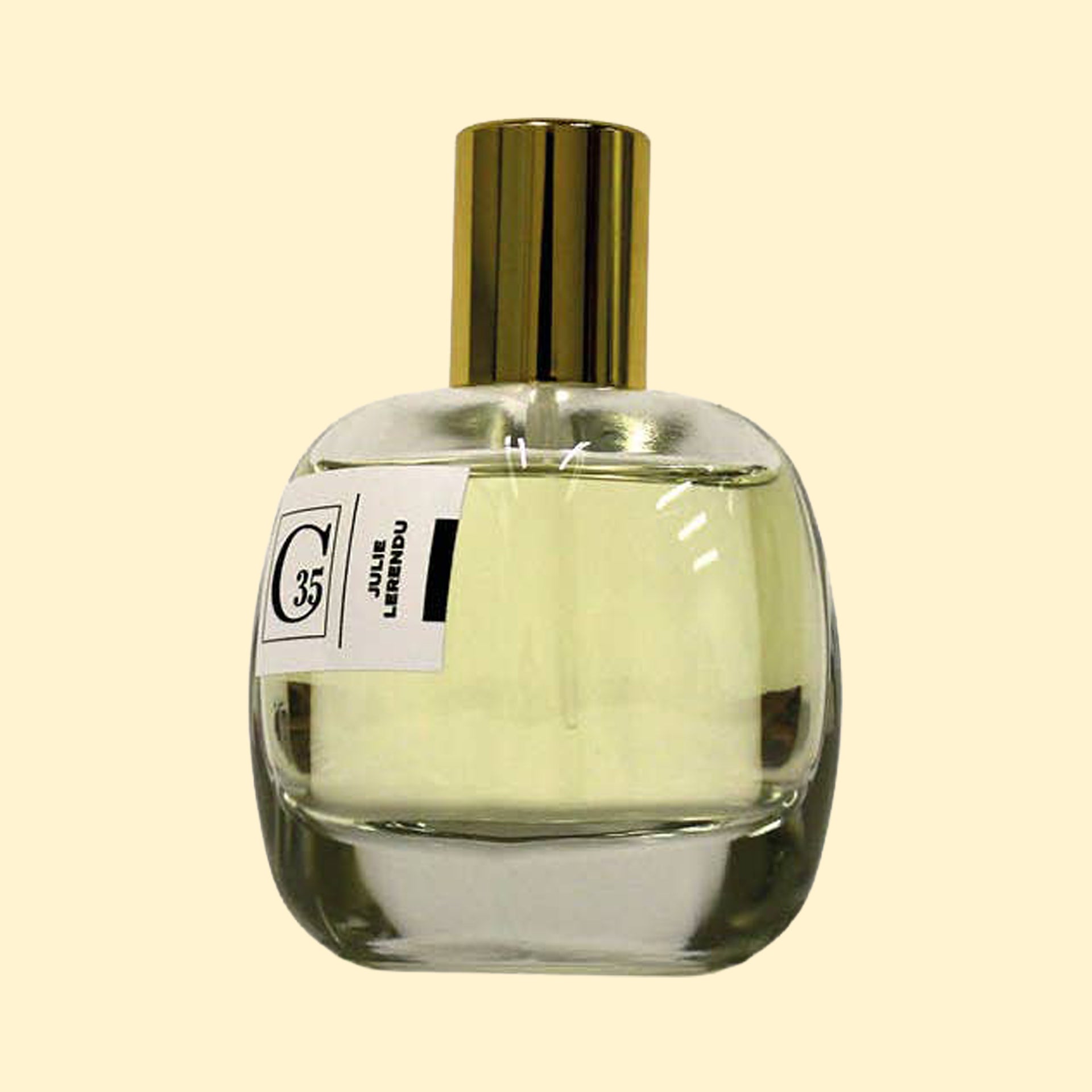 A studio product photograph of a clear glass perfume bottle filled with pale yellow-green fragrance, topped with a reflective gold cylindrical cap and set against a soft cream background. A white label on the front displays the Collection Trente-Cinq logo and the name “Julie Lerendu.” The transparent bottle highlights the soft hue of the perfume, while gentle reflections on the glass emphasize its rounded, sculptural silhouette.
