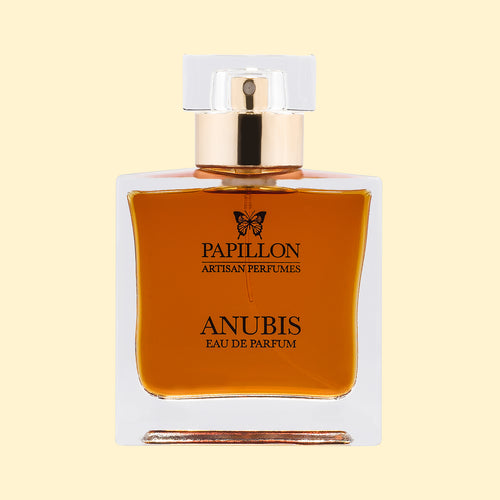 Papillon Artisan Perfumes Anubis Eau de Parfum in a clear rectangular glass bottle with a white cap and gold collar, filled with deep amber-brown fragrance against a soft cream background.